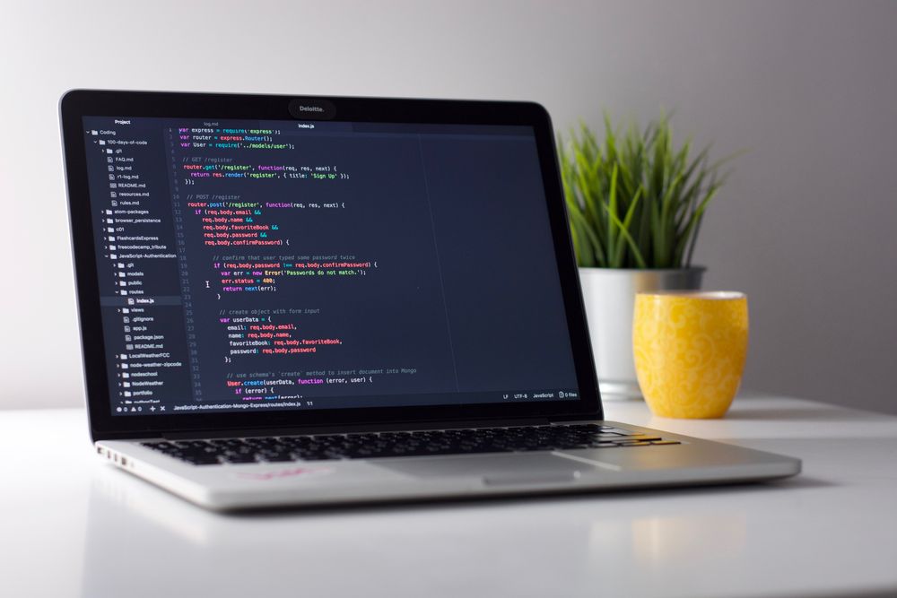 A MacBook with code on its screen sitting on a white desk with a green plant in the far right corner.